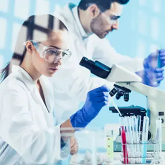 Man and woman in a science lab both wearing protective googles. She is working with  microscope and test tubes filled with various coloured liquids. 