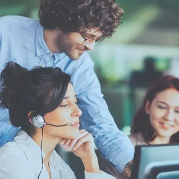 Three people in a customer service centre around a PC monitor, one wearing a phone headset.